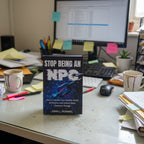 Book titled 'Stop Being an NPC' on a cluttered desk with a computer and coffee cups.